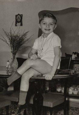 Roma, İtalya 1964 olabilir: Çorap, ayakkabı ve atlet giymiş, arka planda küçük bir dini imge ve bir vazo çiçekli, bir masada oturan gülümseyen çocuk, 1960 'lar veya 1970' ler
