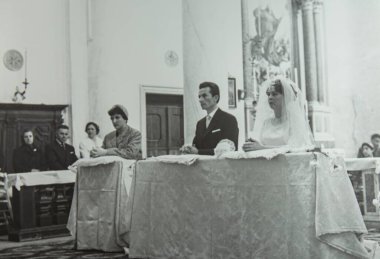 Roma, İtalya 1964 'te olabilir: Bir gelin ve damadın kilisede diz çöktükleri düğün töreninde konuklar ve tanıklarla birlikte çekilmiş klasik siyah beyaz fotoğrafı.