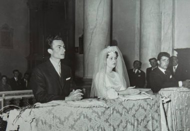 Roma, İtalya 1964 olabilir: Bir gelin ve damadı kilisede diz çökerken gösteren klasik siyah beyaz fotoğraf, konuklarla çevrili.