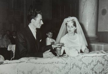 Roma, İtalya 1964: Damat kilisedeki düğün töreninde gelinin parmağına yüzük takıyor. Misafirler ve arka planda bir hizmetçiyle.
