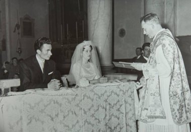 Roma, İtalya 1964 olabilir: Gelin ve damat mihrapta otururken bir rahibin kitap okuduğu bir düğün törenini çeken klasik siyah beyaz fotoğraf