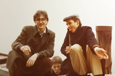 Roma, İtalya 1964: İki genç adam çömeliyor, biri sigara içiyor ve diğeri dilini dışarı çıkarıyor, 1970 'lerden kalma klasik bir fotoğrafta bir çocuk gizlice arkalarından dışarı bakıyor