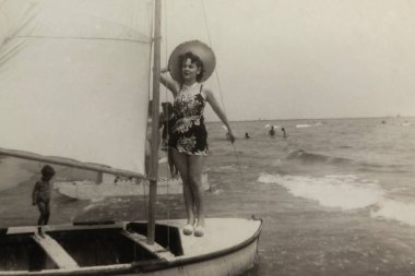 Roma, İtalya 1964: Kıyıya yakın küçük bir yelkenlide poz veren, mayo ve geniş kenarlı bir şapka takan gülümseyen bir kadının fotoğrafı.