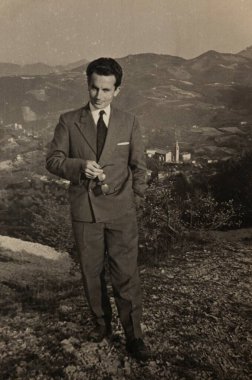 Roma, İtalya 1964: Güneş gözlüğü tutan işadamının portresi, arka planda küçük bir kasaba bulunan pitoresk bir dağ manzarasının önünde klasik bir takım elbiseyle poz veriyor