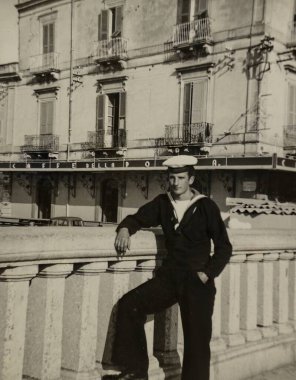 Roma, İtalya 1965 olabilir: 1960 'lardan kalma siyah ve beyaz bir nostaljik fotoğrafta çekilen geleneksel bir italyan kafesinin önündeki balyalara yaslanan kendinden emin genç denizci