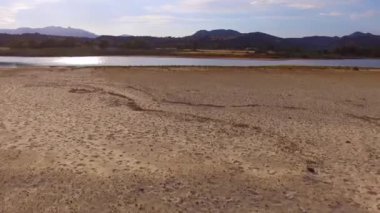 Kum desenleri, parıldayan su ve güneşli bir gökyüzünün altındaki uzak dağların oluşturduğu sakin bir nehir kıyısının havadan görüntüsünü yakalıyorum.