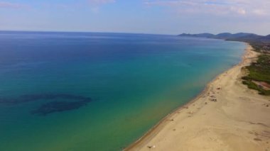 Turkuaz su üzerinde hareket eden kamera, turistlerin ve arka planda bir dağın bulunduğu tropik bir plajı gösteriyor.