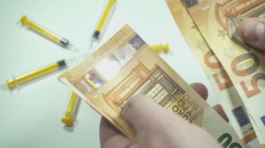 Doktor beyaz arka planda şırıngaların yanında euro banknotları sayıyor.