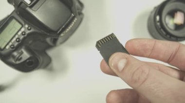 Fotoğrafçı bir hafıza kartı tutuyor, arka planda odak dışı kamera ve lens var.