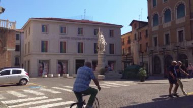 Treviso, İtalya 1 Ocak 2025: Son sınıf öğrencisi bisikletini Treviso, İtalya 'da güneşli bir meydanda sürüyor ve Benetton binasının birleşik renklerinin önünden geçiyor