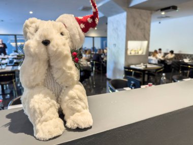Kırmızı Noel Baba şapkalı beyaz pofuduk kutup ayısı oyuncağı gri restoran tezgahında oturuyor, gözlerini kapatıyor ve arka planda bulanık müşteriler yemek yiyor.