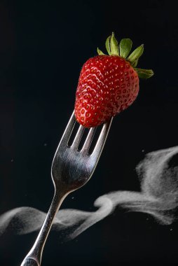 Bekar, olgun kırmızı çilek, gümüş bir çatala nazikçe yerleştirilir, dramatik siyah bir zemin üzerine kurulur, beyaz duman zerreleri bir gizem unsuru ekler.