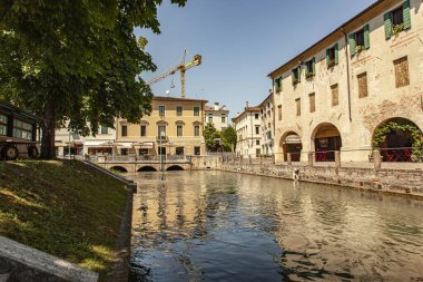 Treviso, Italy 1 January 2025: The cagnan grande canal flowing through treviso, reflecting the colorful buildings and a construction crane in the background, on a sunny summer day clipart