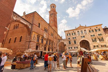 Verona, İtalya 1 Ocak 2025: İtalya 'da pazar tezgahları ve arka planda ortaçağ Torre dei Lamberti ile Verona, İtalya' daki Piazza delle erbe 'yi ziyaret eden turistler