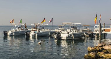 Garda Lake, Italy 3 January 2025: Tourists boarding rental motorboats decorated with various european flags at a dock on lake garda, italy clipart