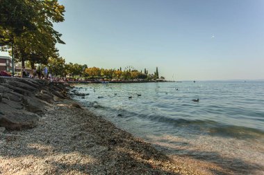 Garda Gölü, İtalya 3 Ocak 2025: Dönme dolap ile Garda Gölü 'nde yüzen ördekler ve güneşli bir günde kıyı boyunca gezinen turistler