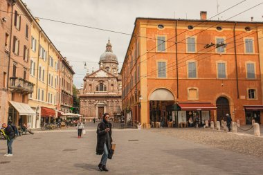 Modena, İtalya 1 Ocak 2025: Arka planda San Biagio kilisesi ile Modena şehir merkezinde yürürken akıllı telefon kullanan ve alışveriş çantası taşıyan şık bir kadın