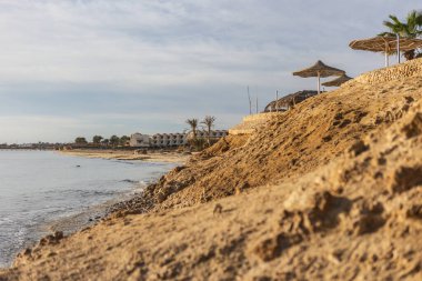 Marsa Alam, Mısır 'daki sakin kıyı şeridi, kumlu kumullar, plaj şemsiyeleri ve bulutlu bir gökyüzünün altındaki uzak bir tatil beldesi.