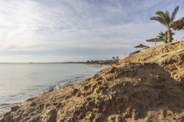 Marsa Alam sahilinde sakin bir sabah, kayalık oluşumlar, palmiye ağaçları, ve yumuşak bir gökyüzü altında uzak oteller