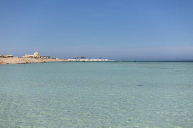 Sakin turkuaz su, Marsa Alam, Mısır 'ın kumlu sahillerini usulca yıkıyor. Sahil şeridinde görülebilen binalar ve palmiye ağaçları, berrak mavi gökyüzünün altında.