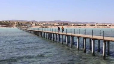 Turistler, turkuaz sulara bakan ahşap iskelede, Marsa alam, Mısır 'daki manzaralı kızıl deniz kıyısındaki tatil beldesinde, tropik cennet manzarasını deneyimleyerek dolaşıyorlar.