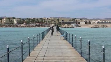 Turkuaz suları boyunca uzanan ahşap iskele, Marsa Alam, Mısır 'da parlak güneş ışığı altında kumlu kıyı şeridine doğru yürüyen turistler.