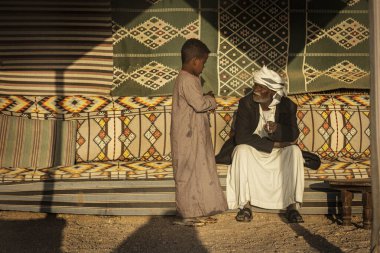 Marsa alam, 9 Şubat 2025: Geleneksel giyinmiş yaşlı bir bedevi, Marsa alam çölünde, Mısır 'da canlı dekore edilmiş bir çadırda genç bir çocukla sohbet ediyor.