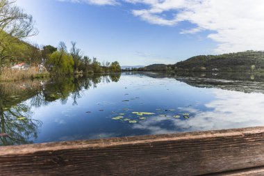 Sakin sular mavi gökyüzünü yansıtıyor, bulutlar ve çevresindeki tepeler Fimon Gölü, İtalya, ahşap bir iskeleden görülüyor.