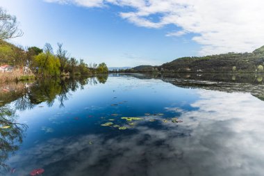 Durgun su gökyüzünü ve bulutları yansıtıyor. Fimon Gölü 'nde. Arkasında nilüferler ve tepeler, güneşli bir günde.