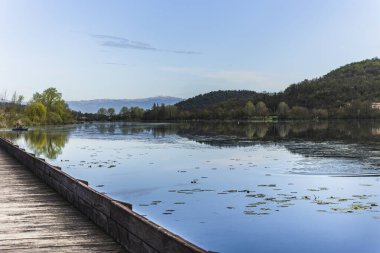 Sakin sular Fimon Gölü 'nün etrafındaki manzarayı yansıtıyor. Sahneye uzanan ahşap iskele ve uzak dağlar sükuneti artırıyor.