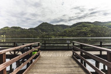 Fimon Gölü 'nün sakin sularına uzanan ahşap iskele, yemyeşil tepeler ve bulutlu gökyüzü tarafından kucaklanarak Veneto, İtalya' da pitoresk bir manzara yaratır.