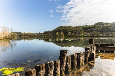 Tahta direkler ve bitki örtüsü fimon gölünün sakin sularına, tepelere ve güneşli bir günde arka planda açık bir gökyüzüne yansıyor.