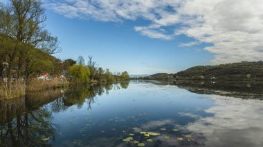 İtalya 'nın Veneto şehrinde güneşli bir bahar günü, fimon gölünün sakin manzarası ağaçları ve gökyüzünü beyaz bulutlarla yansıtıyor.