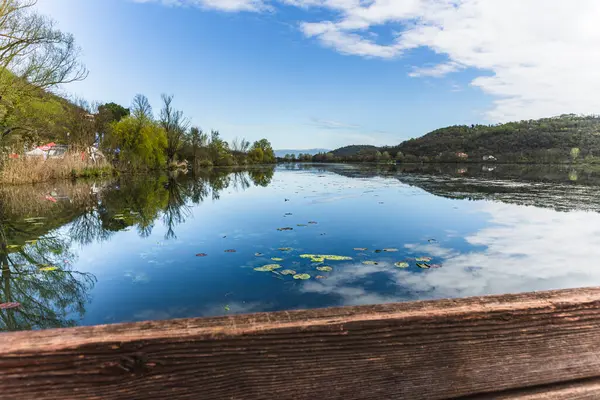 Sakin sular mavi gökyüzünü yansıtıyor, bulutlar ve çevresindeki tepeler Fimon Gölü, İtalya, ahşap bir iskeleden görülüyor.