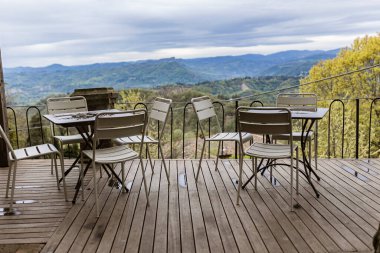 Beyaz metal masalar ve sandalyeler, İtalya kırsalındaki güzel bir vadiye bakan ahşap bir güvertede otururlar.