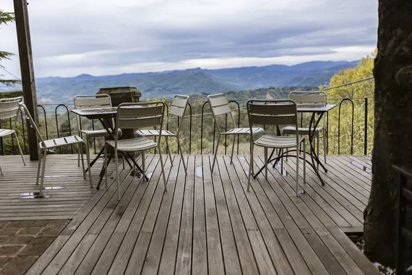 Beyaz metal masalar ve sandalyeler, bulutlu bir günde geniş bir dağ vadisine bakan ahşap bir güvertede otururlar.