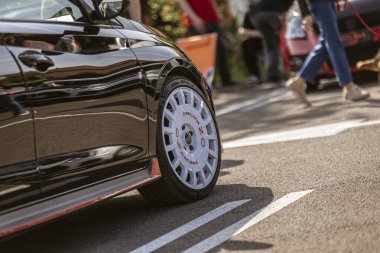 Roma, İtalya, 4 Mayıs 2025: Düşük açı, arka planda bulanık izleyicilerin olduğu bir etkinliğe park edilmiş gösterişli siyah bir spor arabaya monte edilmiş beyaz oz yarış çemberine odaklanıyor.