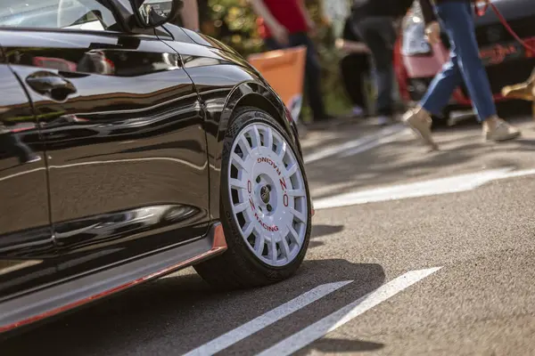 Roma, İtalya, 4 Mayıs 2025: Düşük açı, arka planda bulanık izleyicilerin olduğu bir etkinliğe park edilmiş gösterişli siyah bir spor arabaya monte edilmiş beyaz oz yarış çemberine odaklanıyor.