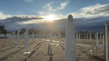 Gün doğumu, kapalı plaj şemsiyelerine ve sottomarinadaki kumlu sahil şeridi boyunca boş sandalyelere yumuşak bir ışık tutarak, huzurlu bir sabah manzarasını gözler önüne seriyor.