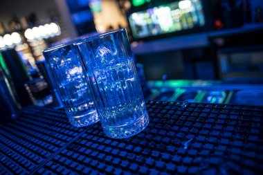 Buzlarla dolu iki bardak boş bir barda mavi ışıkla aydınlatılmış bir bar paspasına oturur.