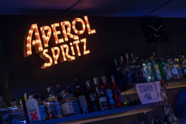 Roma, İtalya, 4 Mayıs 2025: Aydınlatılmış aperol spreyi bir bara sıcak bir parıltı ekler, çeşitli içki şişeleriyle dolu rafların üzerinde, canlı ve davetkar bir atmosfer yaratır.