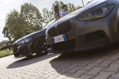 Milan, İtalya 4 Haziran 2025, güneşli bir günde park halindeki iki arabanın yanında duran iki genç, manzaranın tadını çıkarıyorlar.