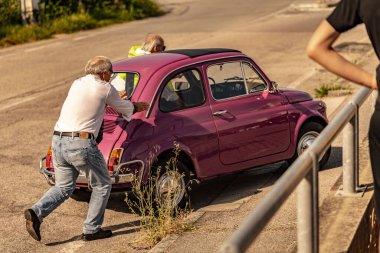 Milan, İtalya, 4 Haziran 2025, son sınıf öğrencisi kırık bir pembe Fiat 500 'ü itiyor. Başka bir son sınıf öğrencisi güneşli bir yolun kenarında oturuyor.