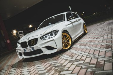 Roma, İtalya, 17 Haziran 2025, şık beyaz BMW M140i sergilenen altın jantlar, geceleyin bir benzin istasyonunda tuğla kaldırıma park edilmiş.