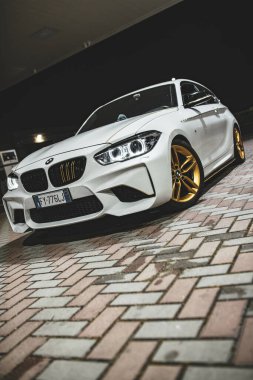 Roma, İtalya, 17 Haziran 2025, şık beyaz BMW M2 spor tasarımı ve altın jantlarını sergiliyor, gece ışıklarının loş parıltısının altında tuğladan bir yüzeye park etmiş.