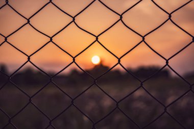 Bulanık bir batan güneş akşam karanlığında gökyüzünde ılık turuncu renkler sergilerken, tel örgülü bir çite odaklanmak.