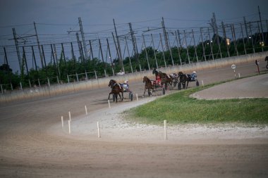 Padua, İtalya 8 Temmuz 2025. Atlar ve sürücüler gece gökyüzünün altında elektrik hatları ve yeşilliklerle çevrili bir toprak pistte koşum koşusunda yarışıyorlar.