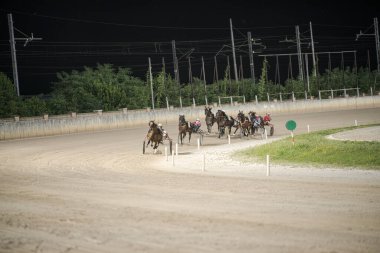 Padua, İtalya 8 Temmuz 2025, açık bir gökyüzü altında yeşil ve elektrik hatlarıyla çevrili kumlu pistte yarışan atlar ve sürücüler.