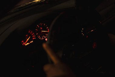 Aydınlatılmış gösterge paneli ve direksiyon simidi gece bir arabanın içinde görünür, dramatik bir atmosfer yaratır.