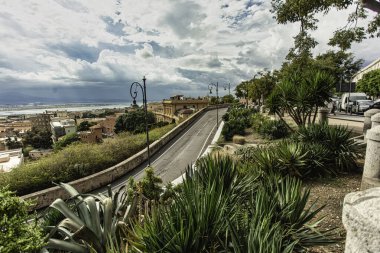 Cagliari, İtalya 19 Ağustos 2025: bereketli bitki örtüsü ve sokak lambalarıyla kaplı dolambaçlı yol, Cagliari 'nin pitoresk kentsel manzarasının panoramik bir manzarasına götürür.